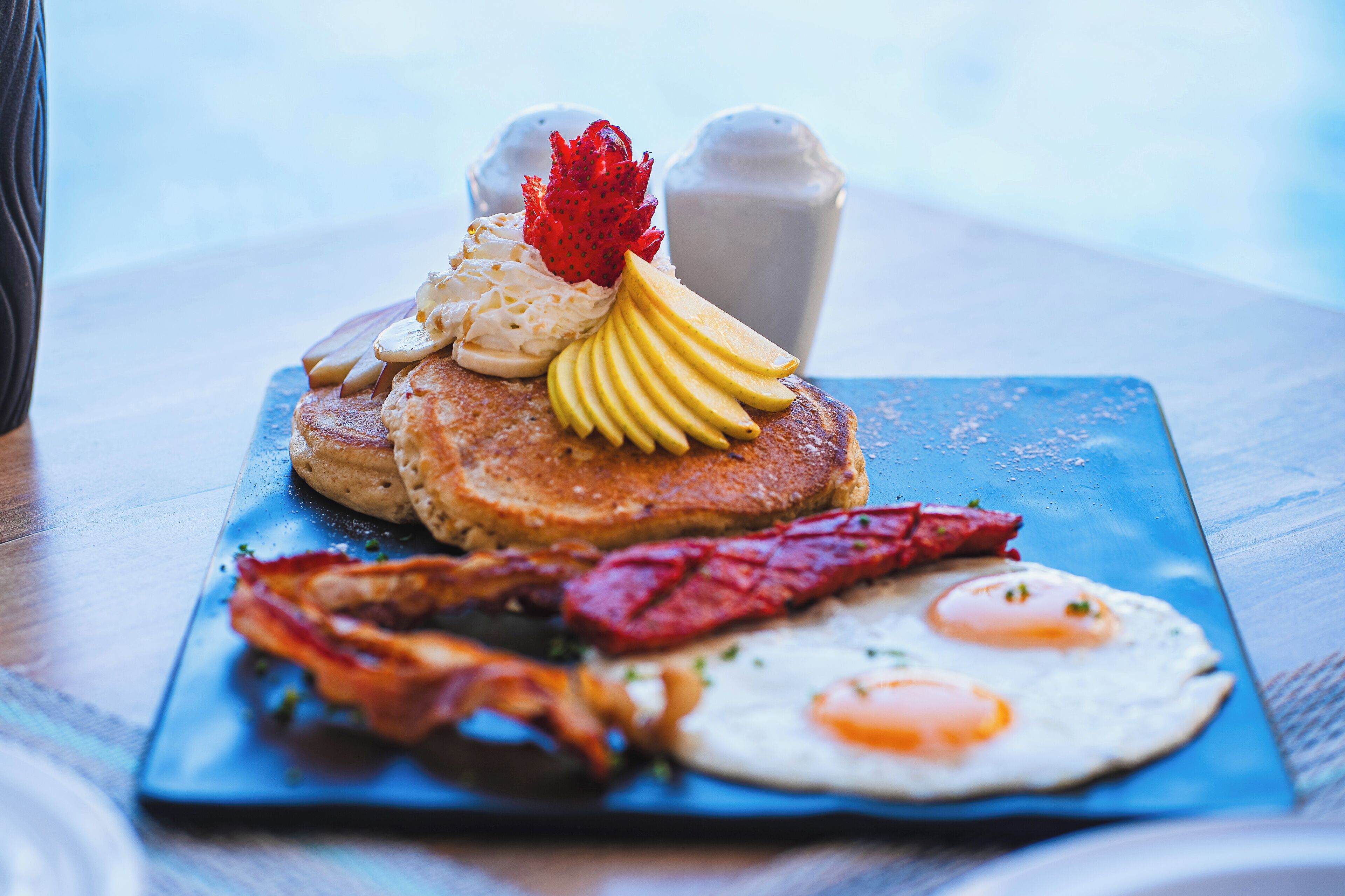 A breakfast plate with pancakes, eggs, bacon, and fruit on a blue plate.
