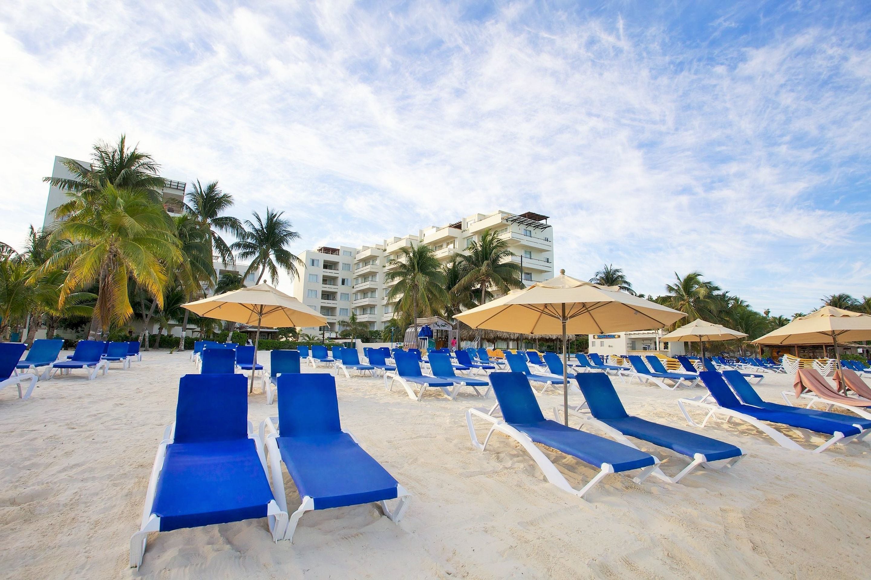 A beach with rows of blue lounge chairs and umbrellas, palm trees, and a hotel building in the background.