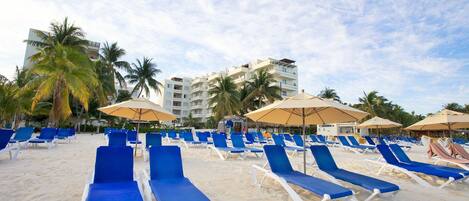 A beach with rows of blue lounge chairs and umbrellas, palm trees, and a hotel building in the background.