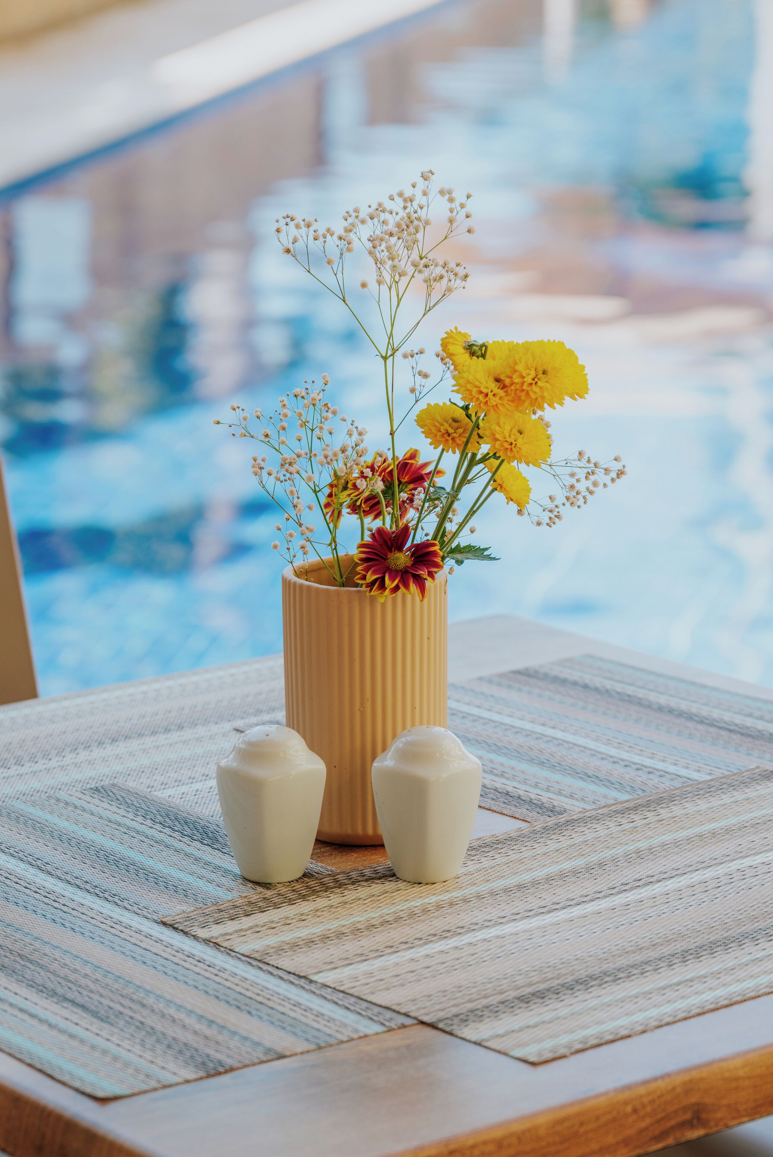Una mesa de madera con un jarrón de flores y dos objetos blancos similares a velas junto a una piscina.