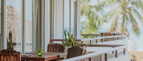 A balcony with wooden furniture and a view of palm trees and the ocean.