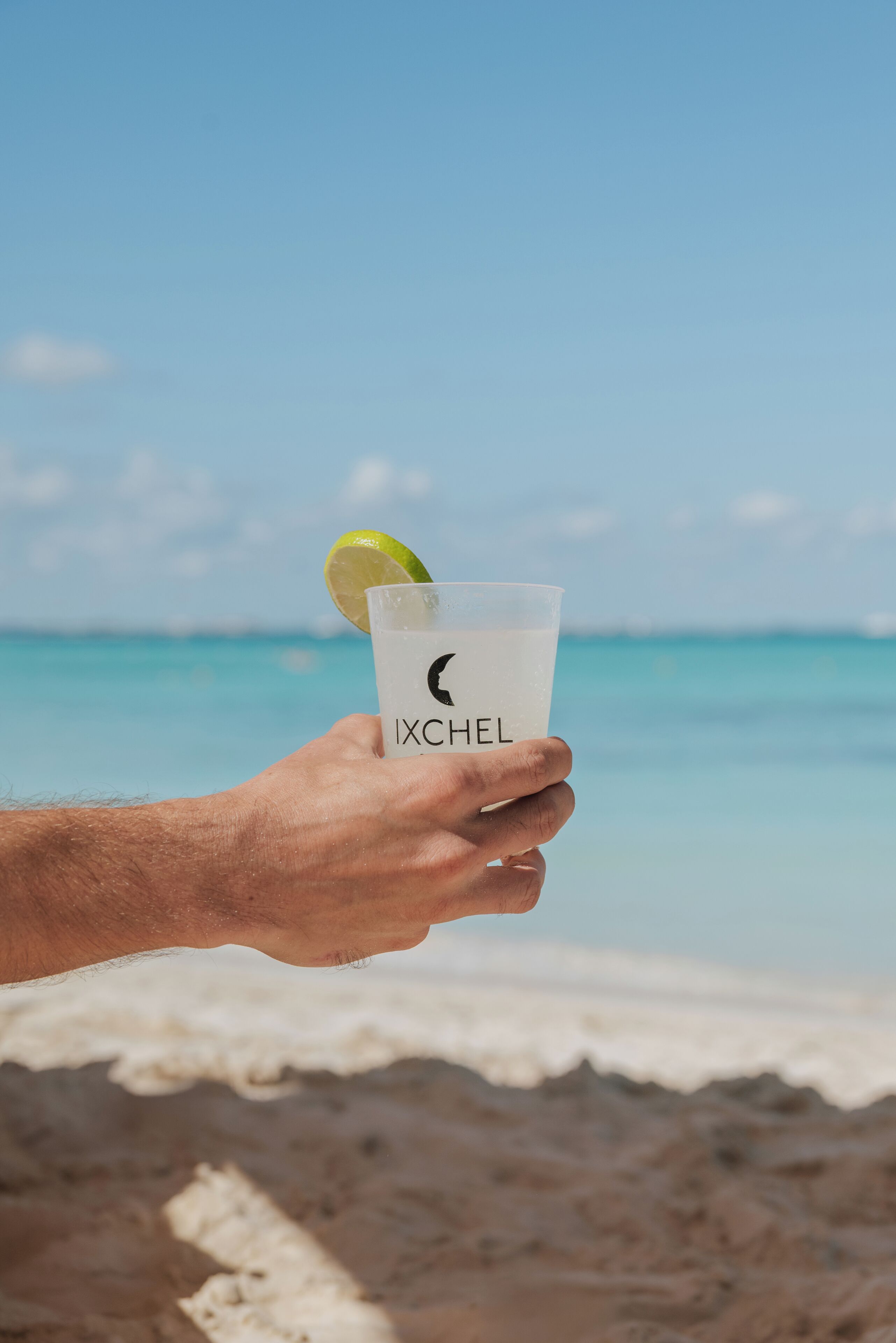 A hand holding a glass with a lime wedge, labeled "IXCHEL," against a beach backdrop.