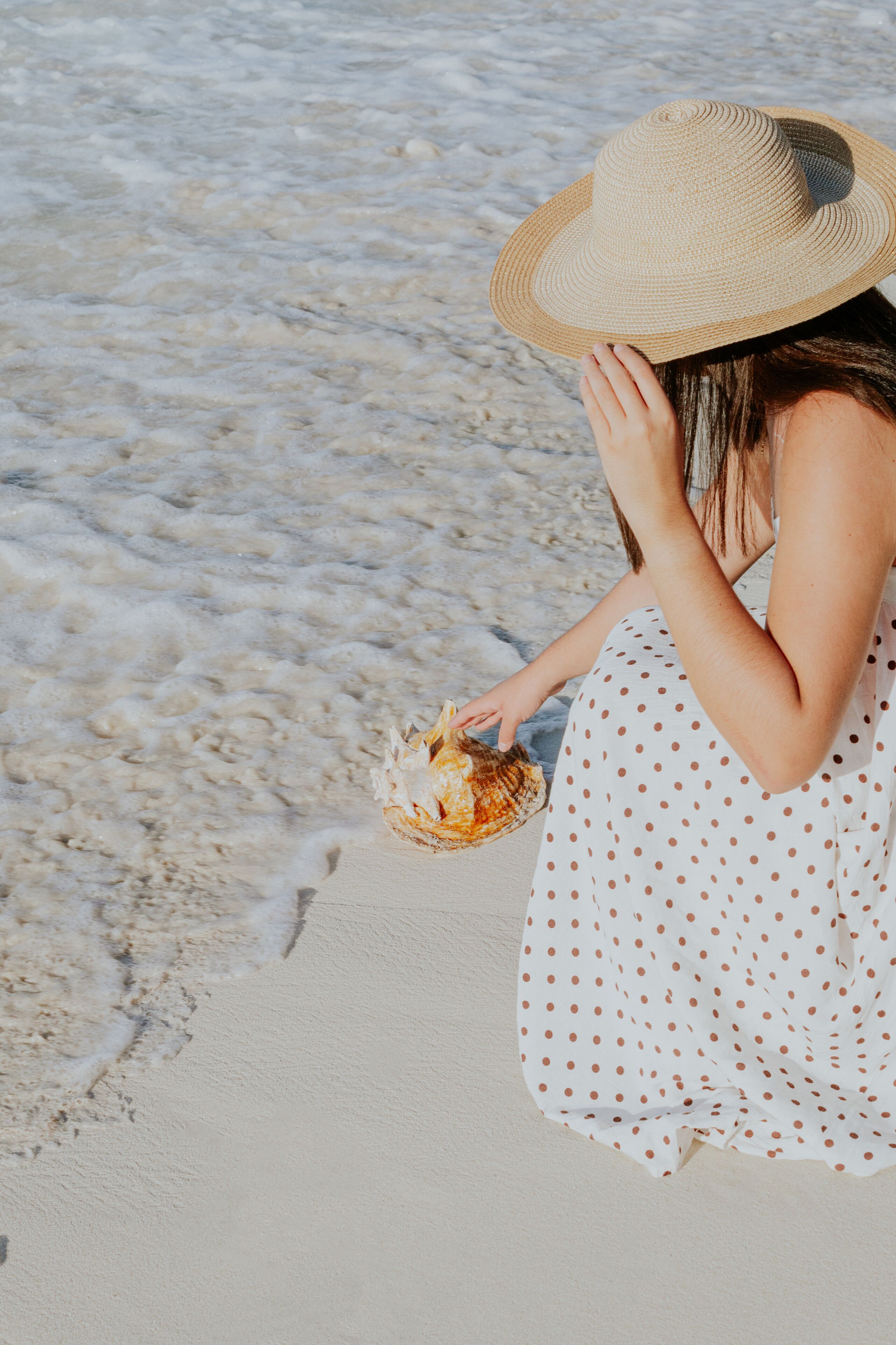 Una persona sentada en la playa, vestida con un vestido de lunares y un sombrero de paja, sosteniendo una concha.