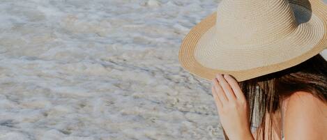Una persona sentada en la playa, vestida con un vestido de lunares y un sombrero de paja, sosteniendo una concha.