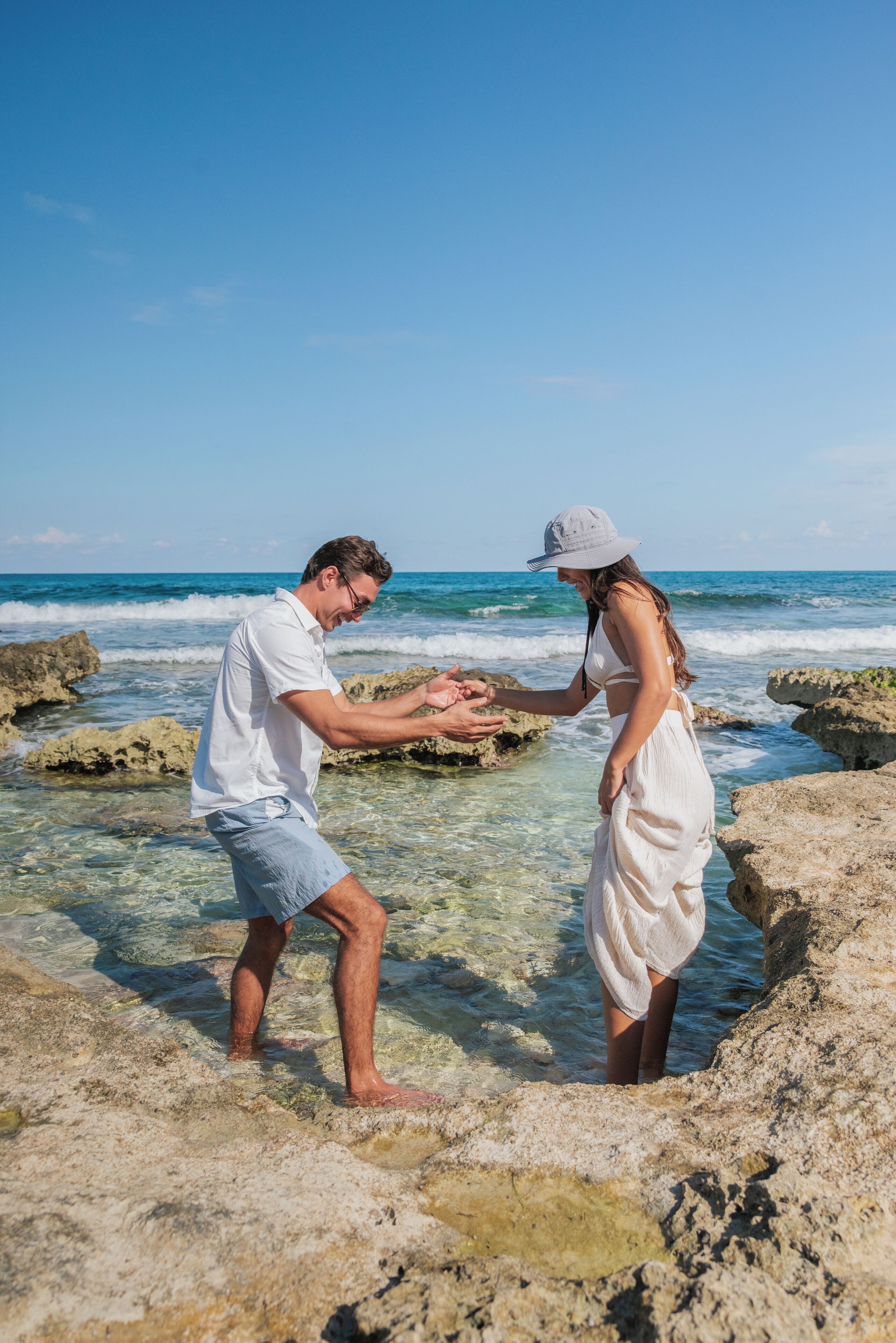 Un couple se tenant la main sur une plage rocheuse, avec une eau claire et des vagues de l’océan en arrière-plan.