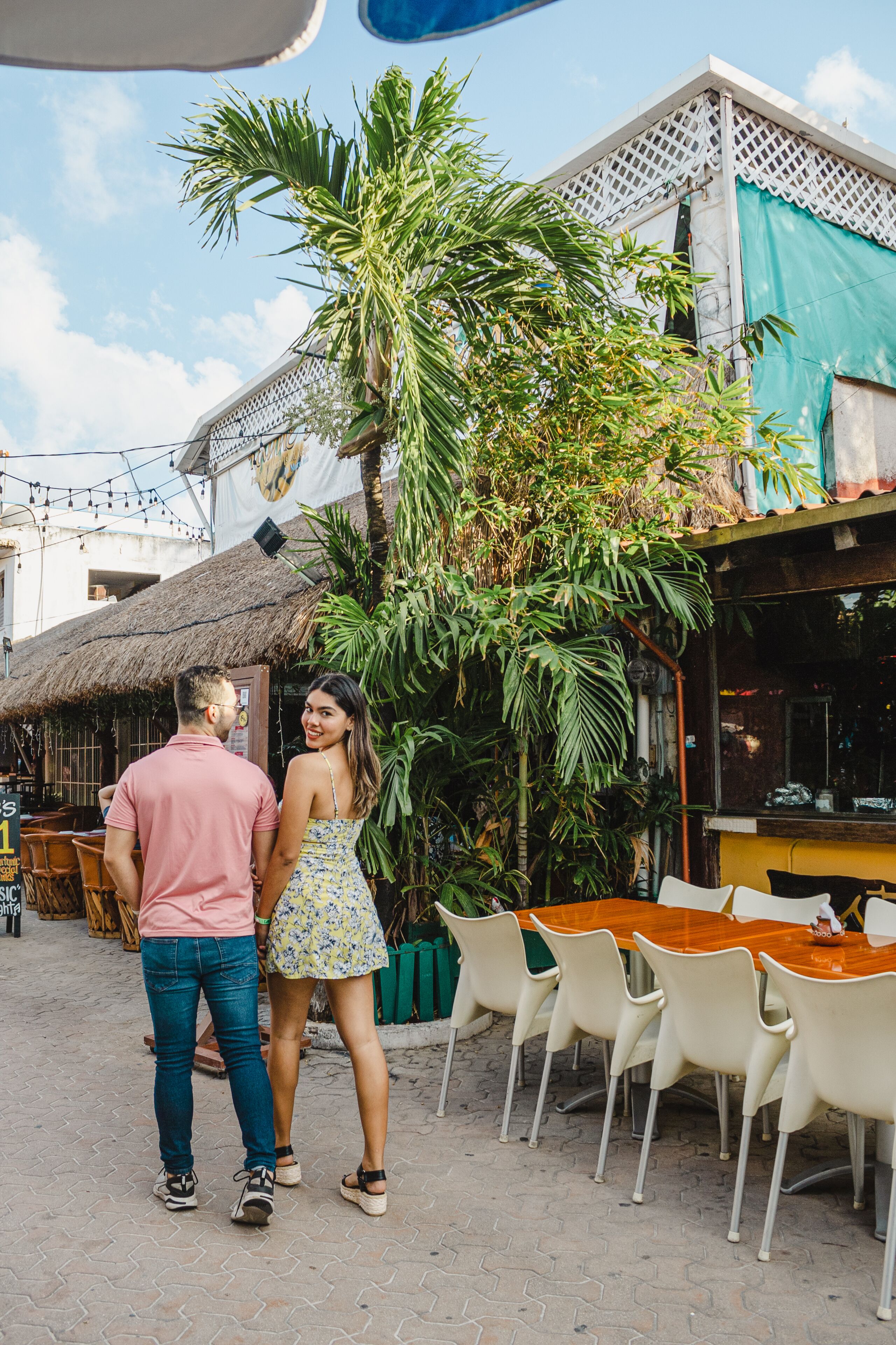 Una pareja parada frente a un restaurante con terraza al aire libre, rodeada de follaje y un edificio con tejado de paja.