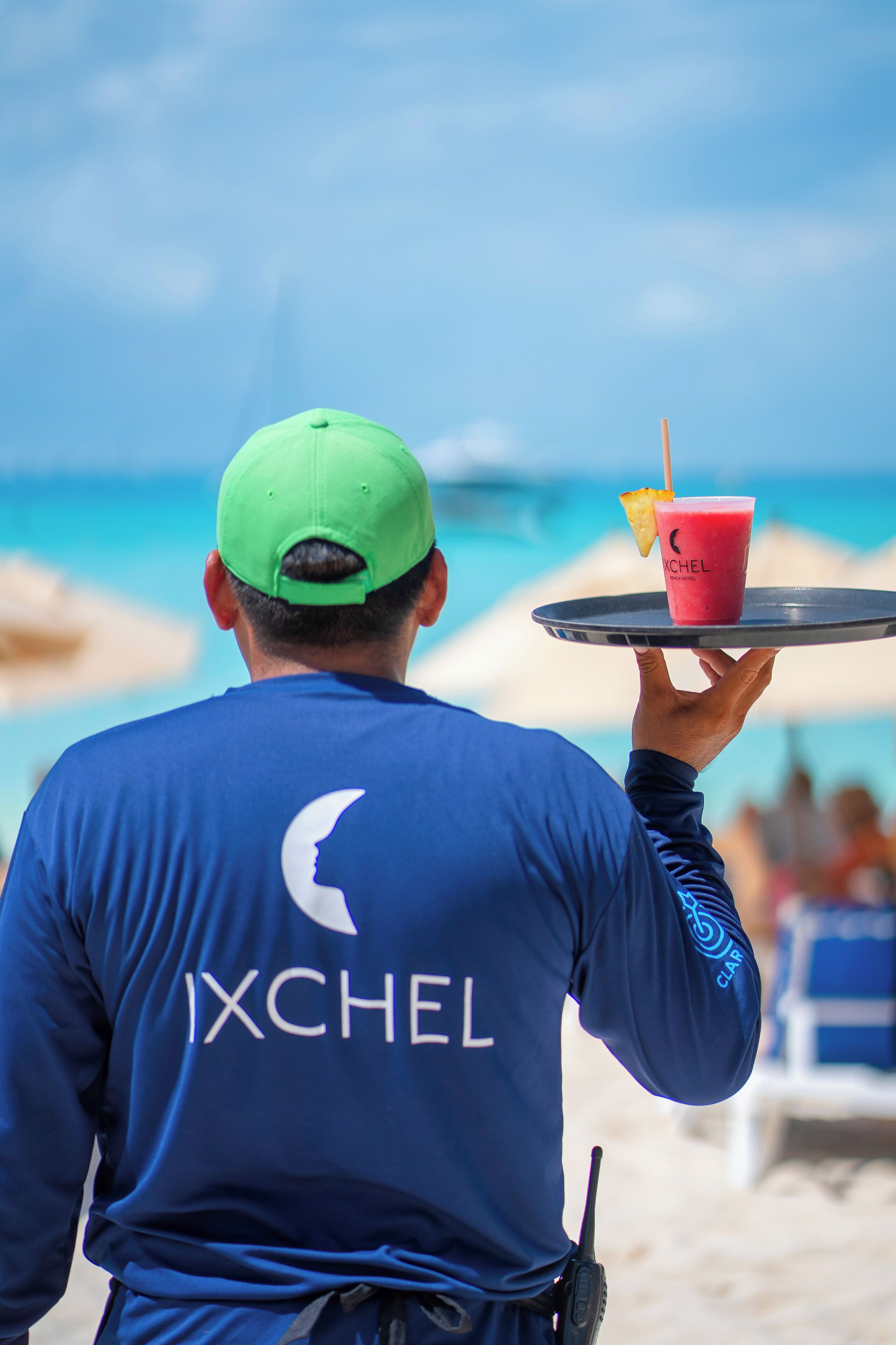 A person in a blue shirt with "IXCHEL" on the back, holding a tray with a drink and a lemon wedge.