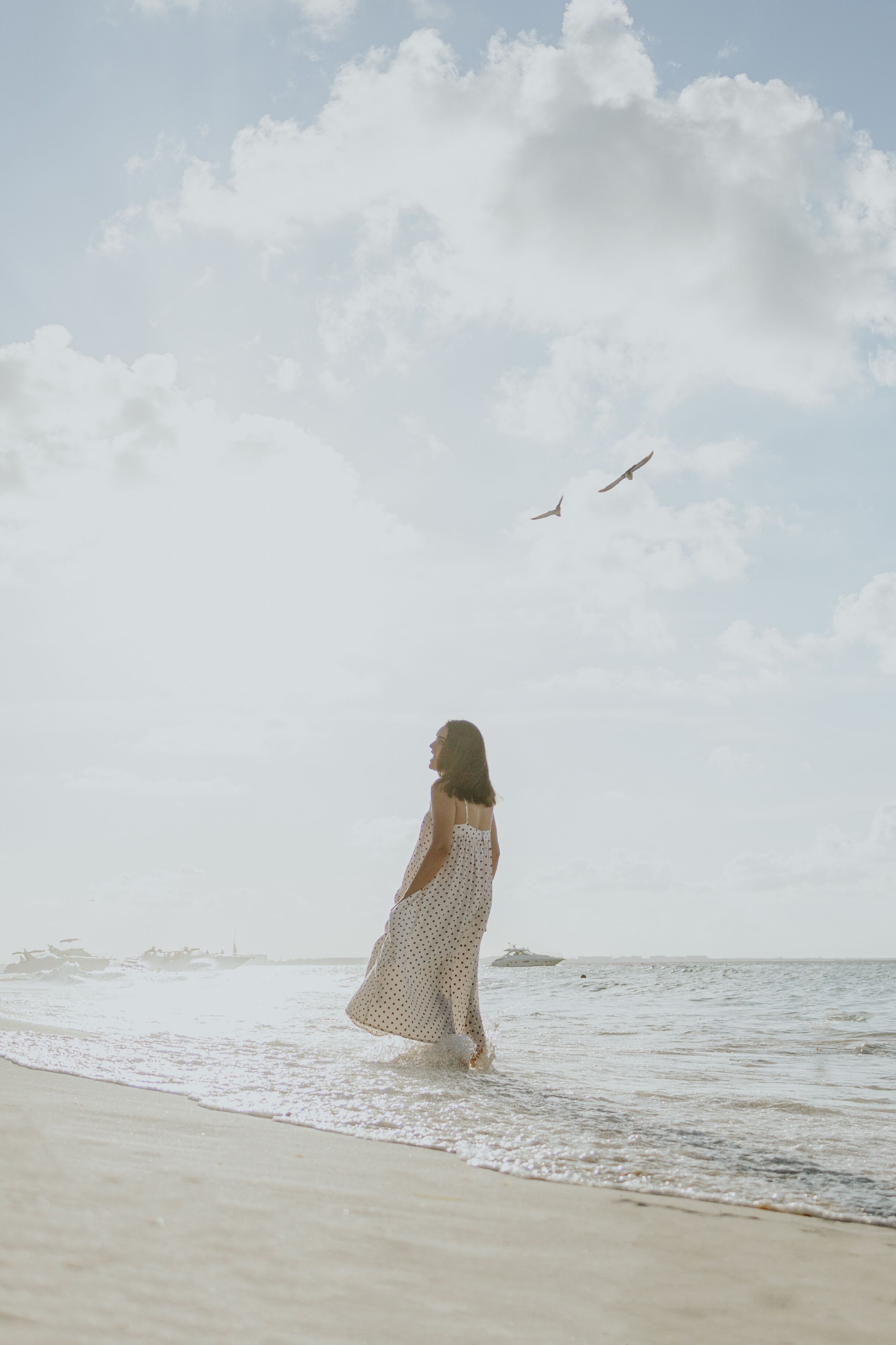 A woman in a polka dot dress standing on a beach with the ocean and birds in the sky.