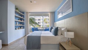 A modern hotel room with a large bed, a bedside table with a lamp, a balcony with a view of palm trees and a building, and a shelf with decorative items.