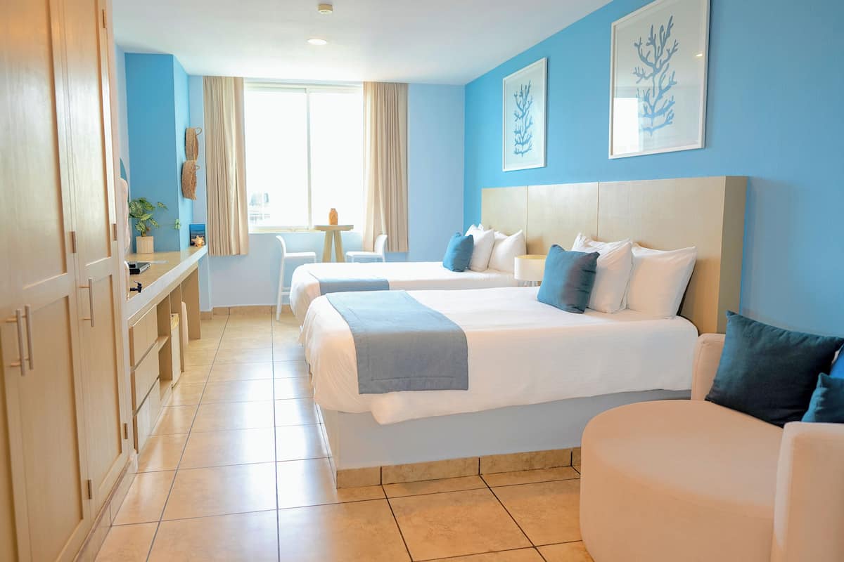 A hotel room with two beds, a desk, and a chair. There is a window with curtains, a plant, and artwork on the wall.