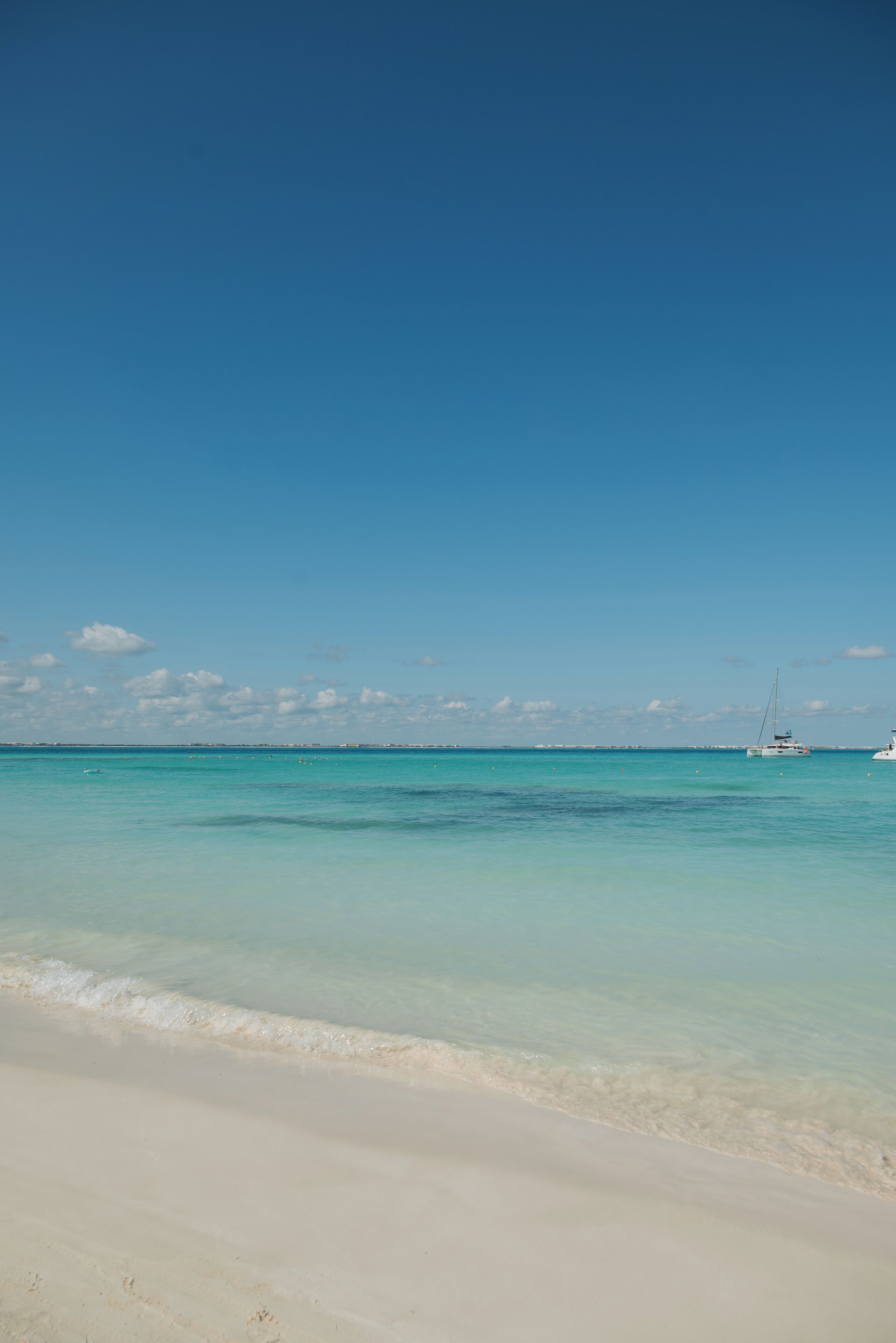 A beach with clear turquoise water and a sailboat in the distance.