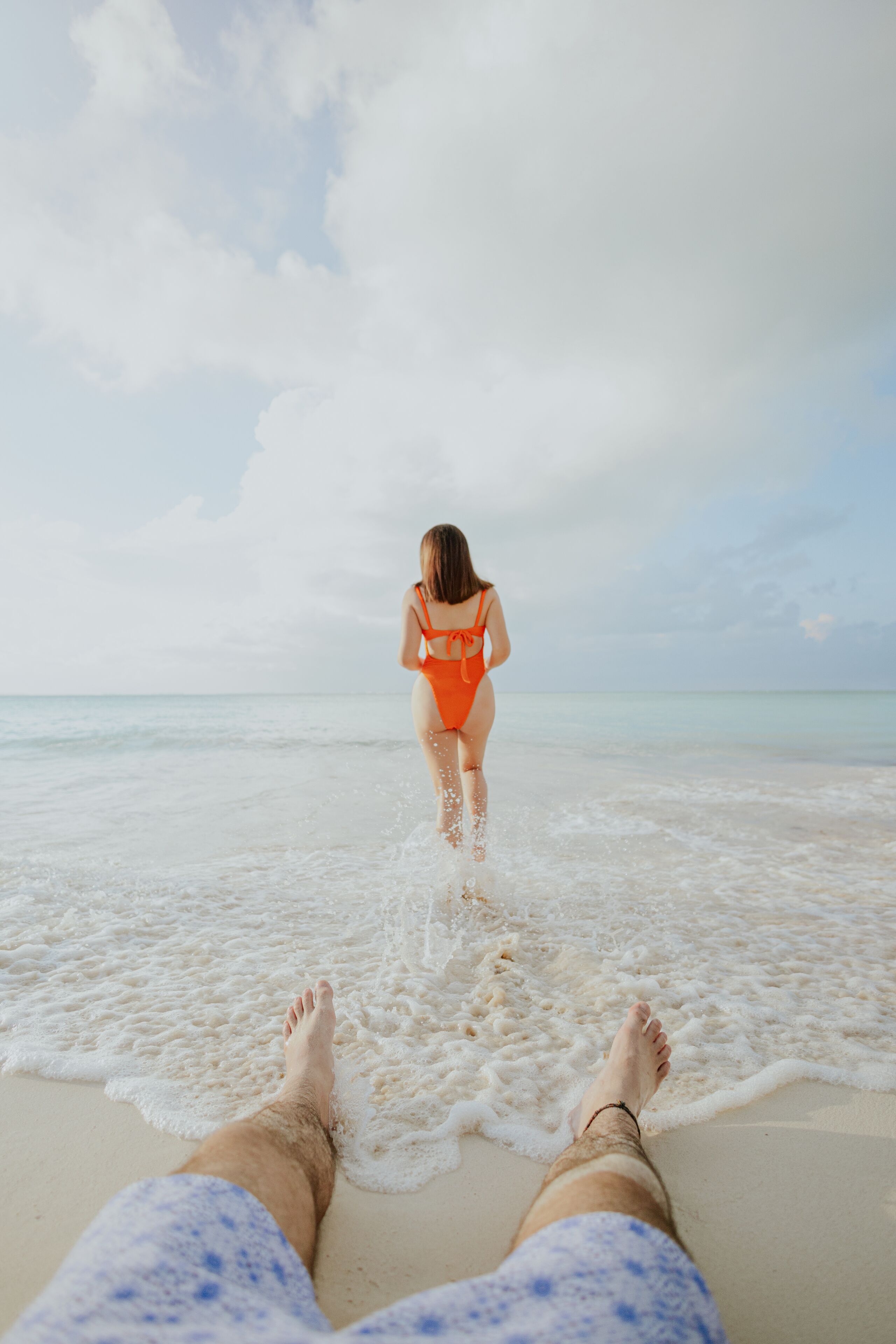 A beach scene with a person in an orange swimsuit walking towards the water, and another person lying on the sand with their feet visible.