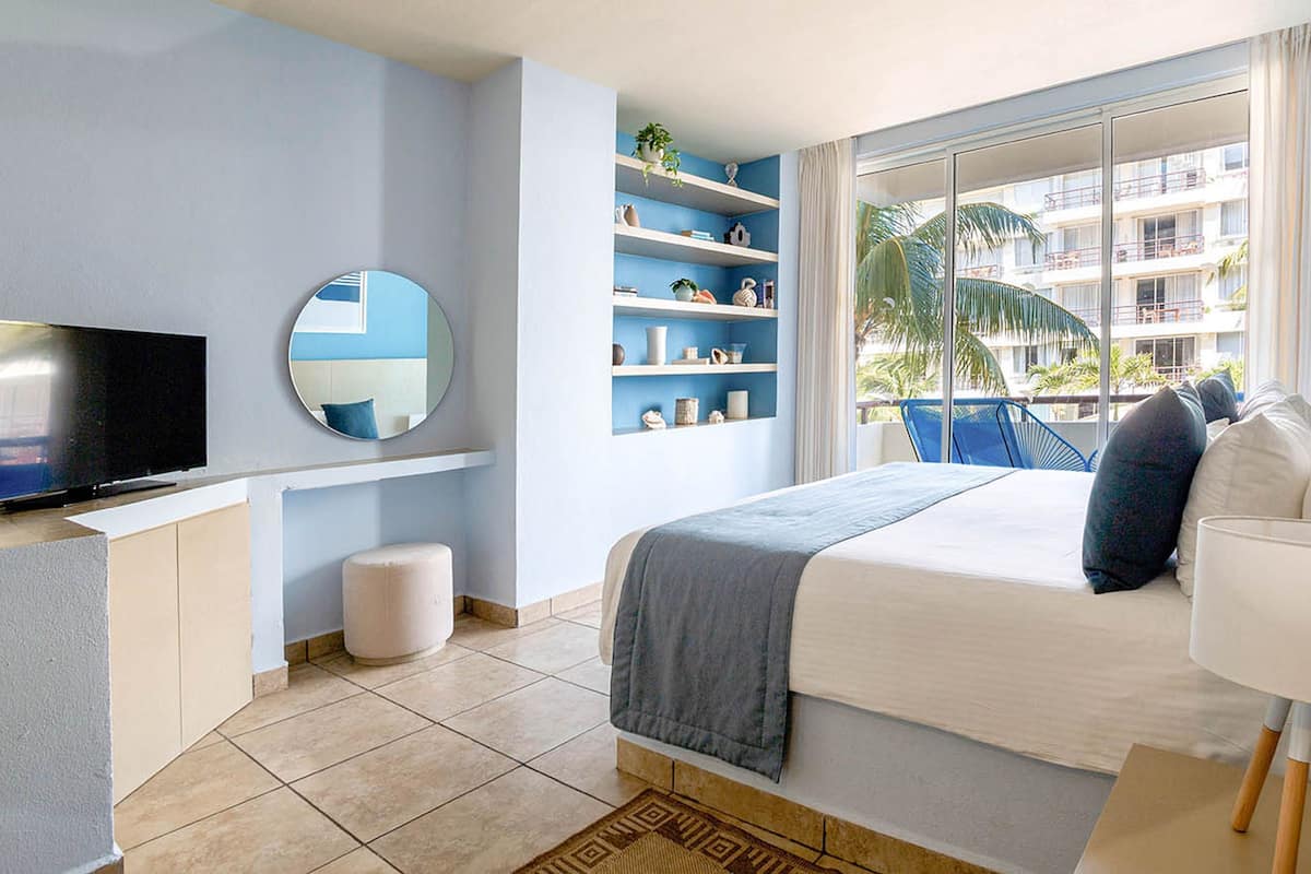A modern hotel room with a large bed, a TV, a round mirror, and a balcony with a view of the building.