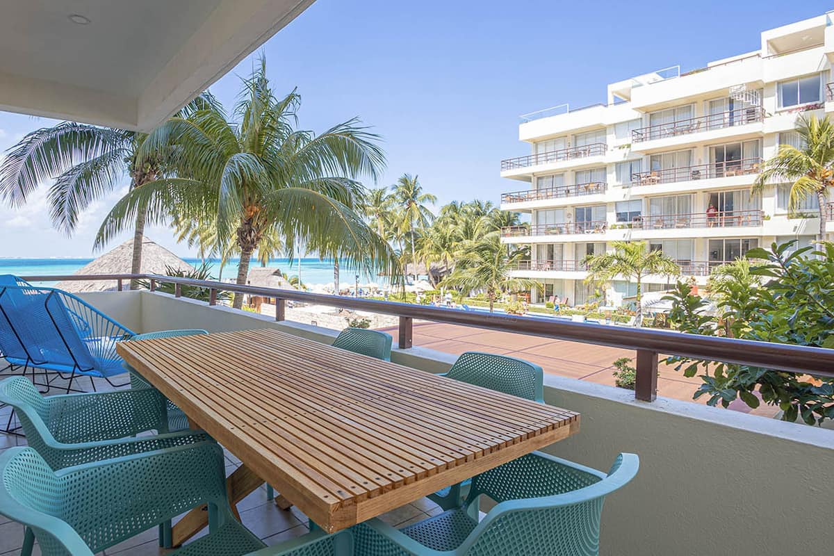 A balcony with a wooden table and green chairs, overlooking a beach with palm trees and a hotel building.