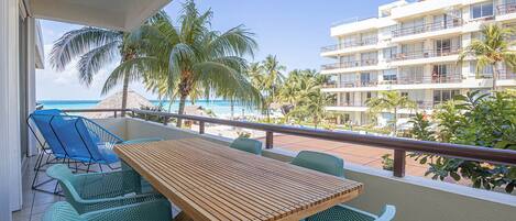 Un balcón con una mesa de madera y sillas verdes, con vista a una playa con palmeras y un edificio de hotel.