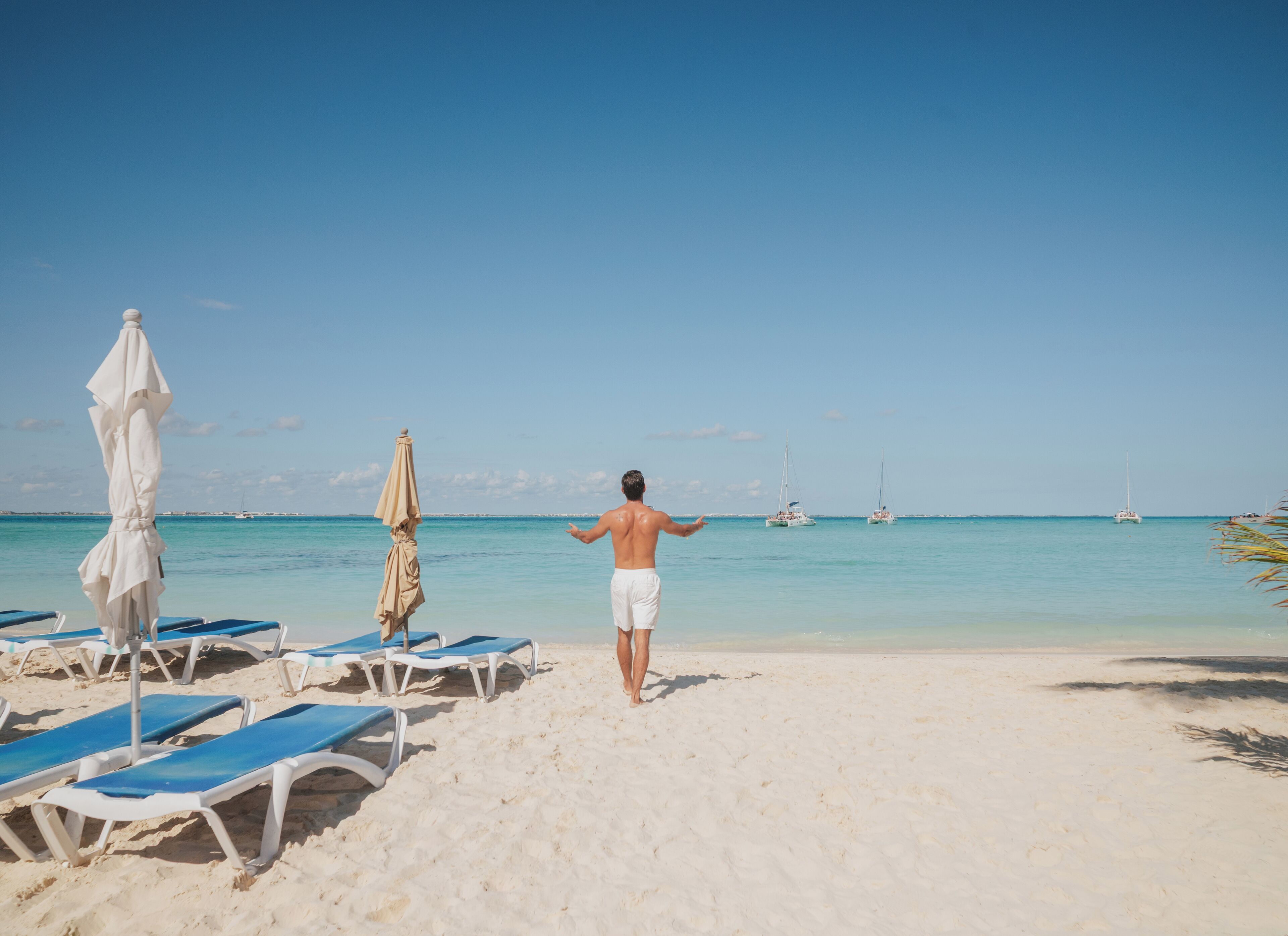 A beach with sun loungers and umbrellas, a clear sky, and sailboats in the distance.
