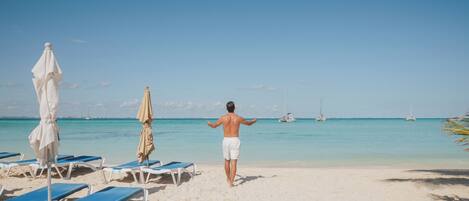 A beach with sun loungers and umbrellas, a clear sky, and sailboats in the distance.