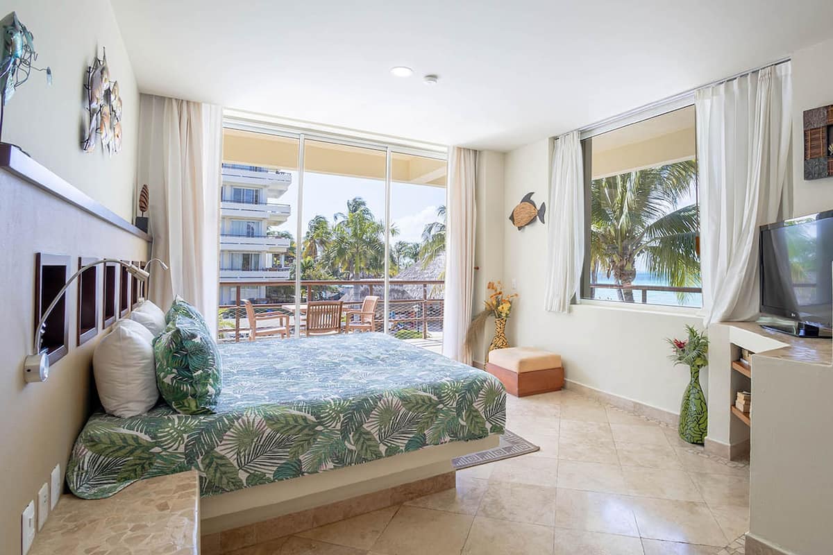 A hotel room with a bed, a TV, a balcony with a view of palm trees, and a dining area with chairs.