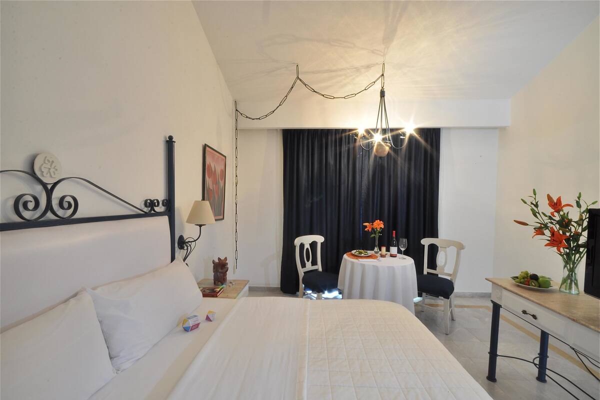 A hotel room with a bed, a desk, a chair, a table with a vase of flowers, and a painting on the wall.