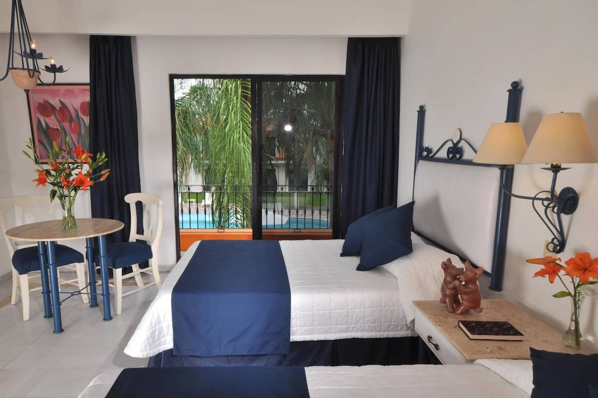 A hotel room with a bed, a desk with a chair, a small table with a vase of flowers, a lamp, and a view of a pool and greenery outside.