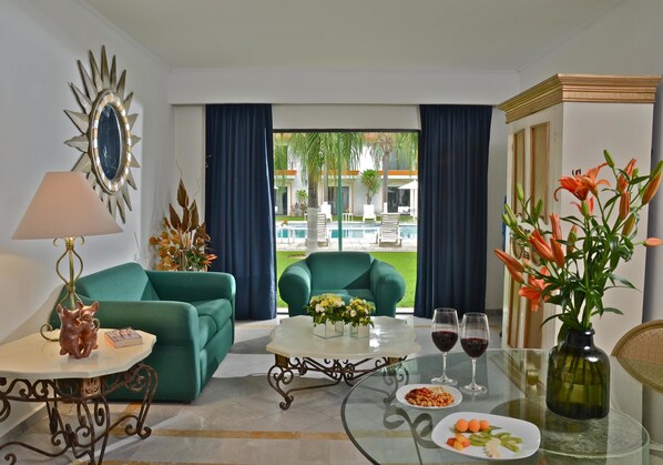 Habitación de hotel con sofá verde, una mesa de centro de cristal con flores, una mesa de comedor con platos y copas de vino, y vistas a una piscina y palmeras afuera.