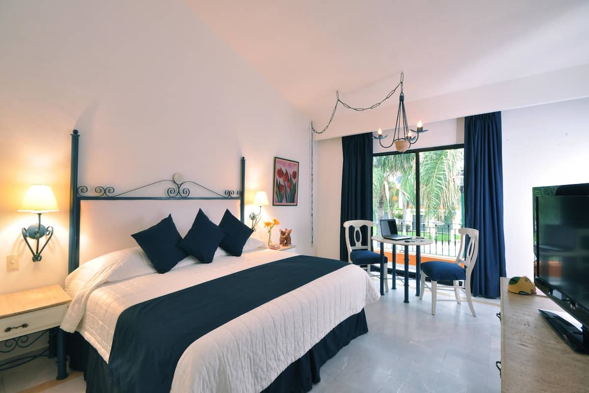 A hotel room with a large bed, a desk with a chair, a TV, and a balcony with a table and chairs.
