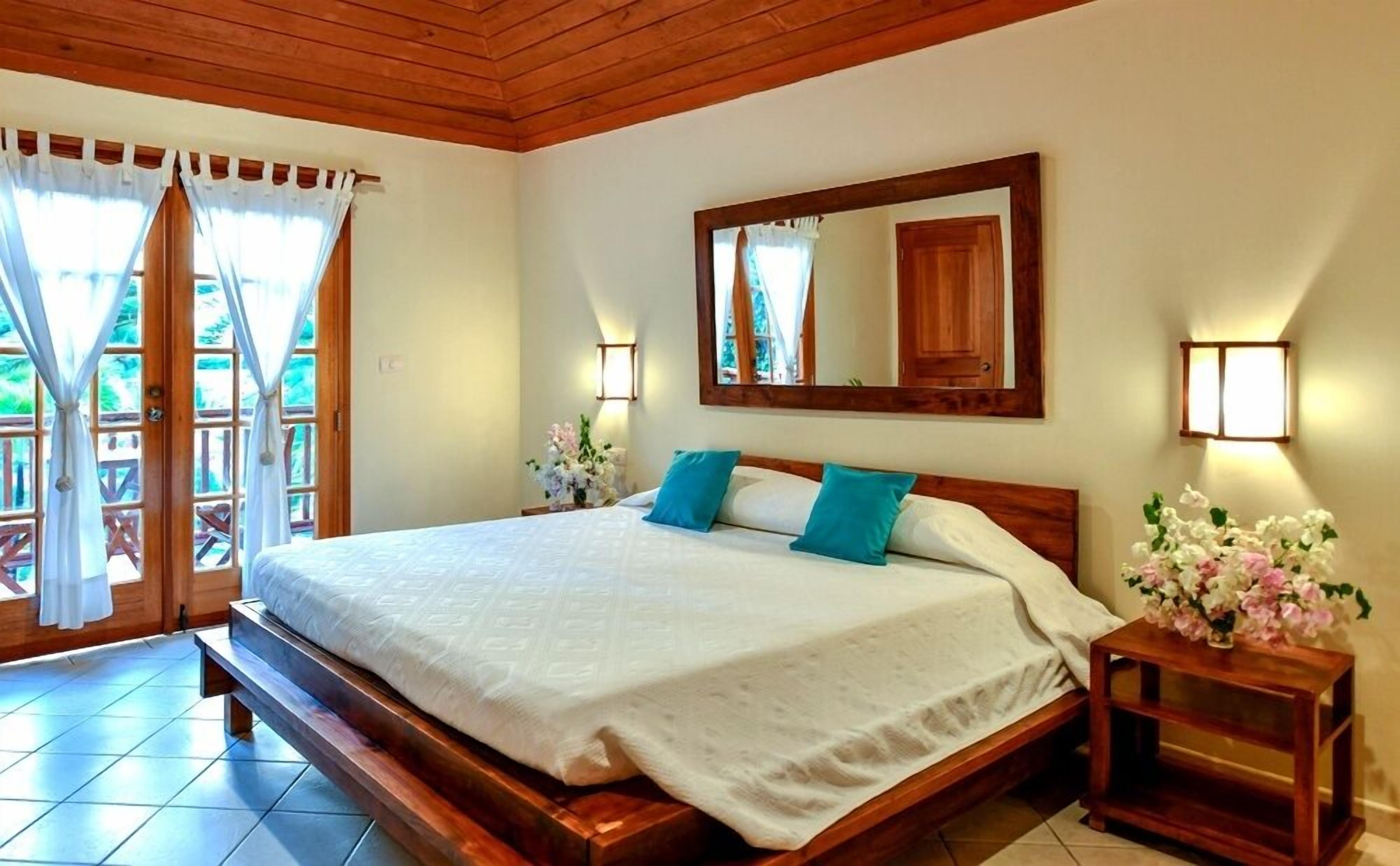 A bedroom with a wooden bed, white linens, teal pillows, a large mirror, and a nightstand with a vase of flowers.