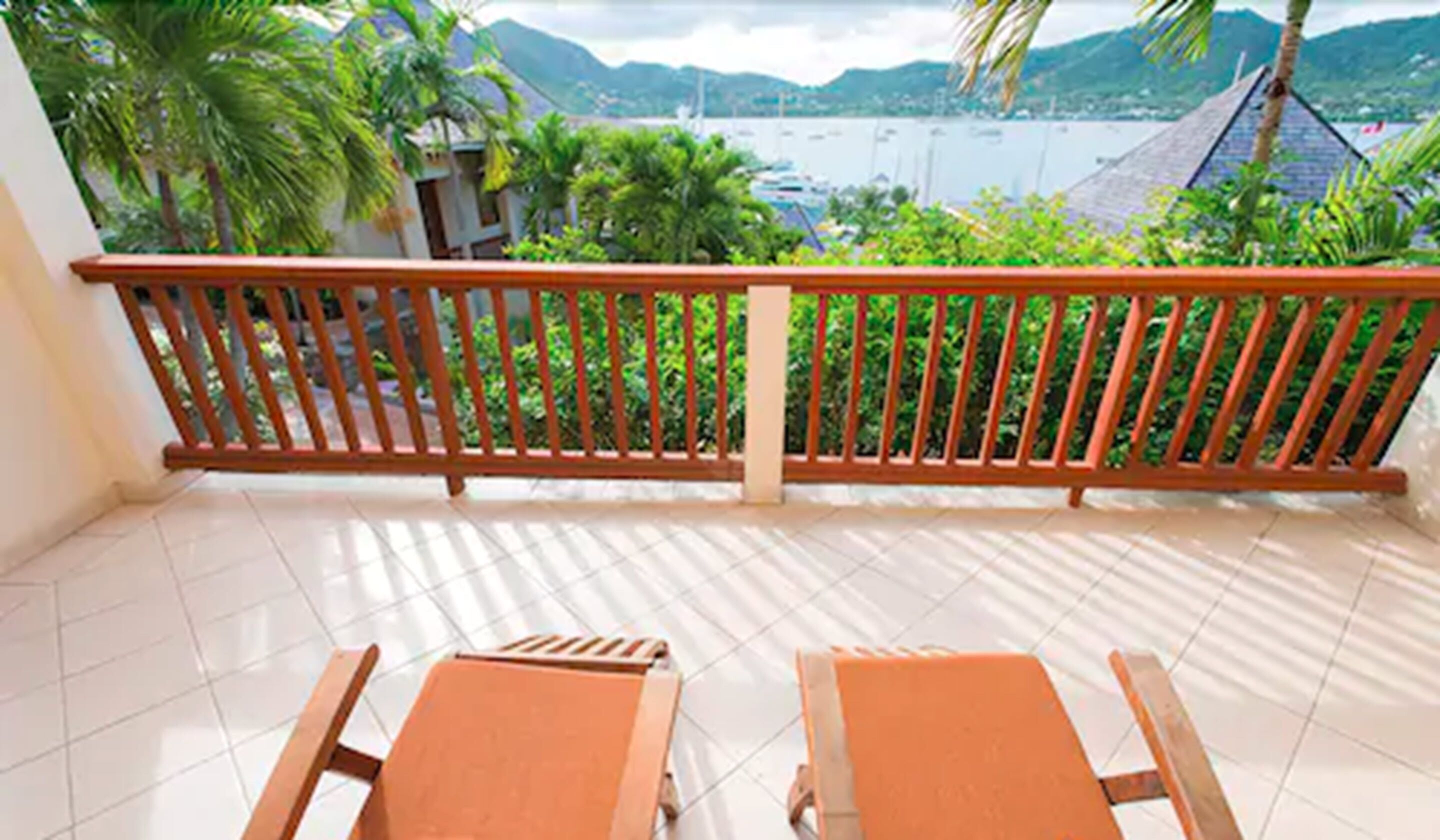 A balcony with a wooden railing, two wooden chairs, and a view of a body of water with mountains in the background.