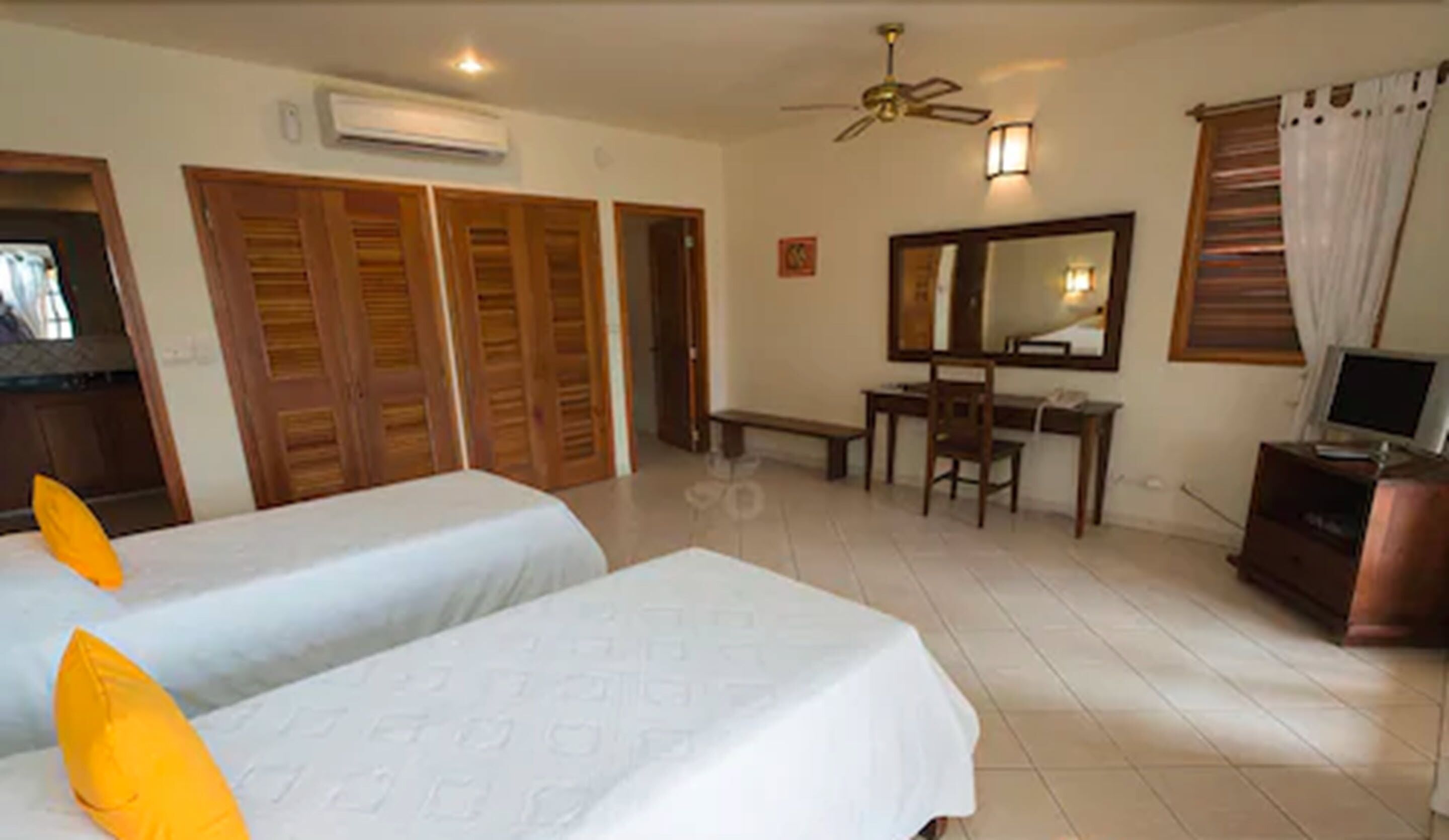 Habitación de hotel con dos camas, un escritorio, una silla, televisión, ventilador de techo y un espejo grande.