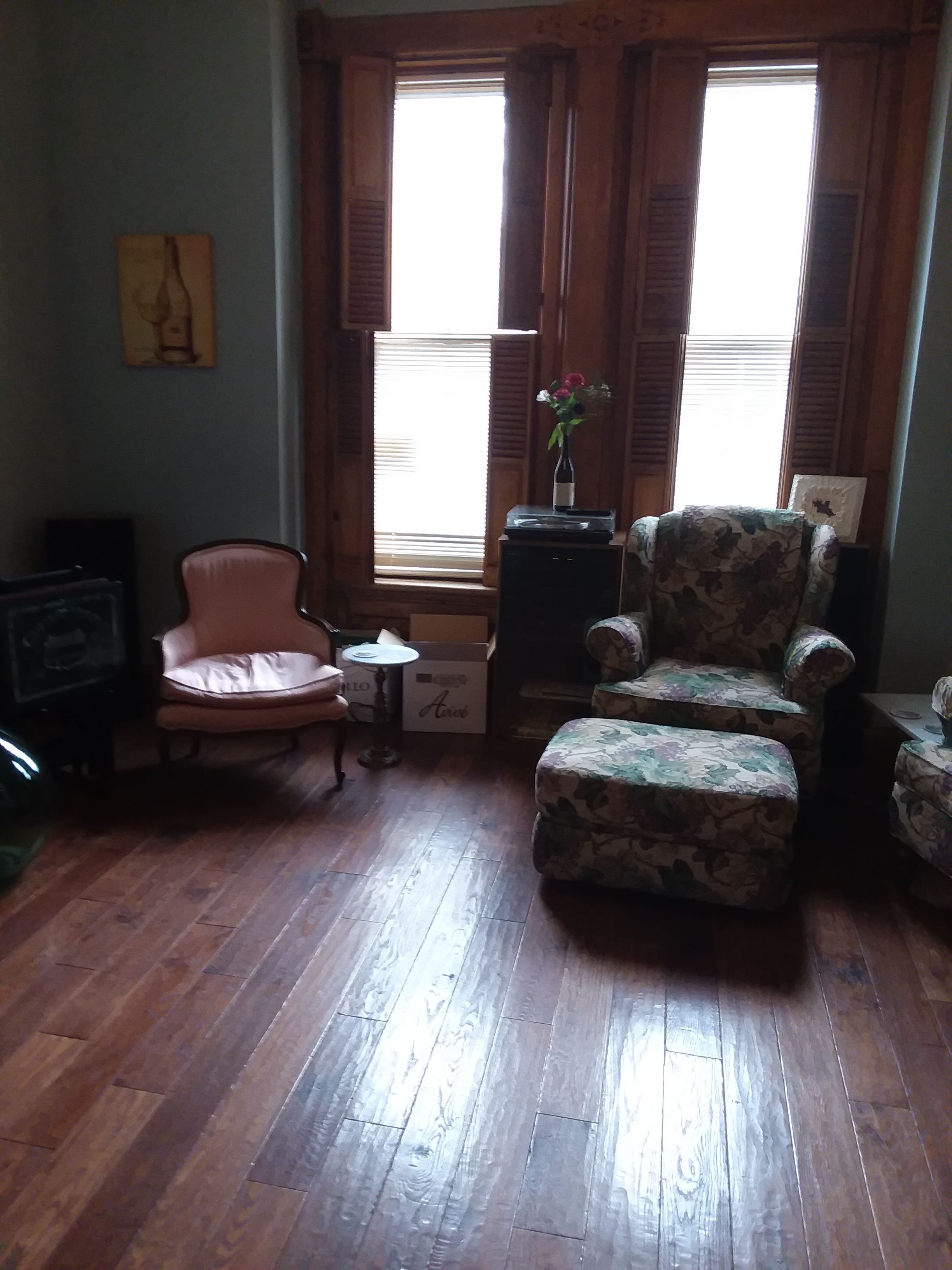 A cozy living room with wooden flooring, a floral armchair, a vintage armchair, and a small table with a vase of flowers.