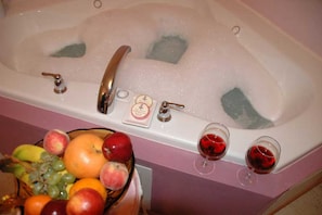 A bathtub with bubbles, a fruit bowl, and two glasses of red wine.