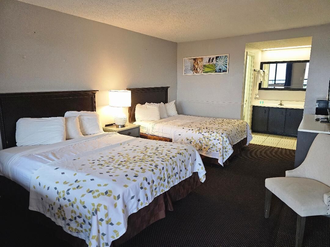 A hotel room with two beds, a desk, a chair, a nightstand with a lamp, and a bathroom visible through an open door.