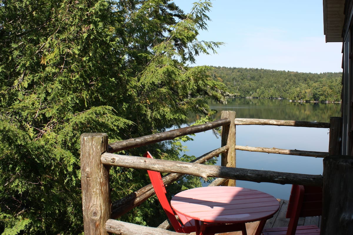 Une terrasse en bois avec une chaise rouge et une table, d’où l’on a vue sur un lac, le tout entouré d’une forêt dense.