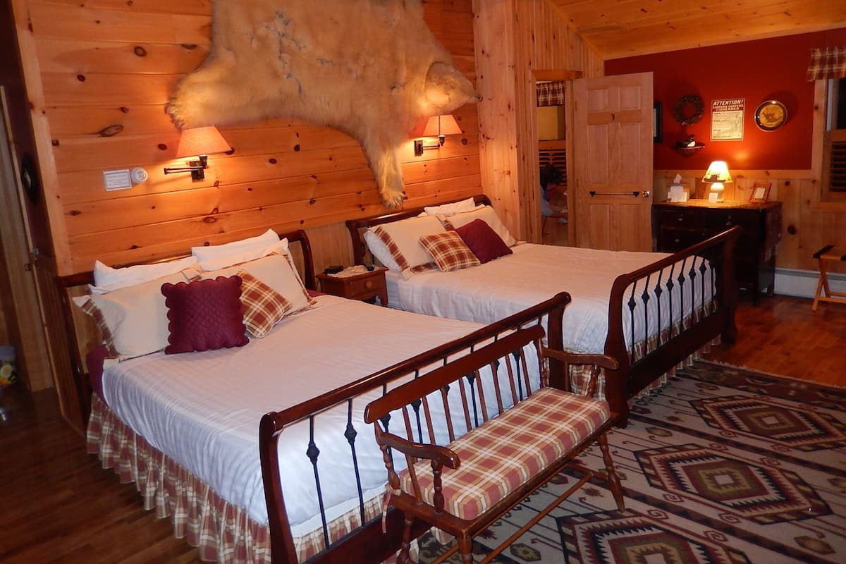 Une pièce aménagée dans le style d’un chalet en rondins, avec deux lits en bois, un banc, une tête de lit en bois ornée d’un morceau de fourrure, un mur rouge et un tapis à motifs.