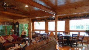 A cozy cabin interior with a wooden ceiling, a large sofa, a dining table with chairs, and a large window offering a view of the outdoors. - Kiwassa Lake Bed & Breakfast (Saranac Lake)