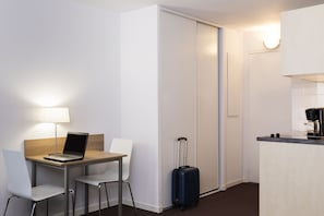 Quarto de hotel com escrivaninha, laptop, mala, cadeira e cozinha compacta.