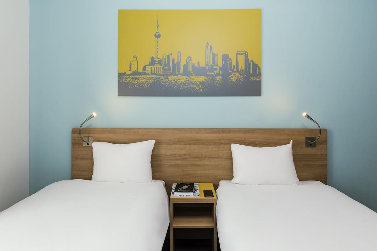 Une chambre d’hôtel avec un lit, deux oreillers, une table de chevet avec un livre et un téléphone, et un tableau représentant un paysage urbain accroché au mur.