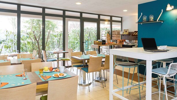 A modern dining area with large windows, a white counter with a laptop, and various seating arrangements.