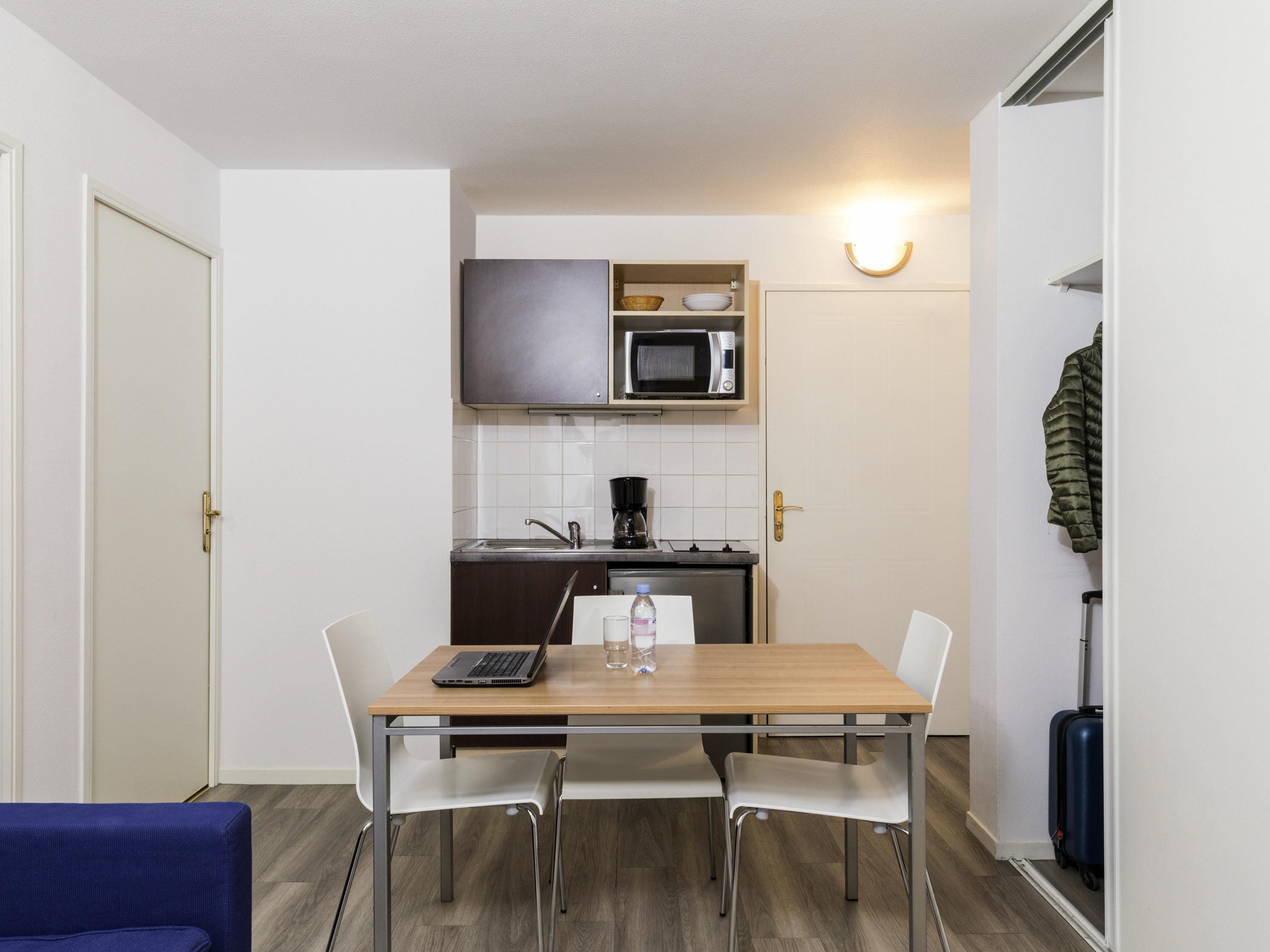A compact kitchen with a microwave, coffee maker, and a small cabinet. There is a dining table with two chairs and a laptop on it.