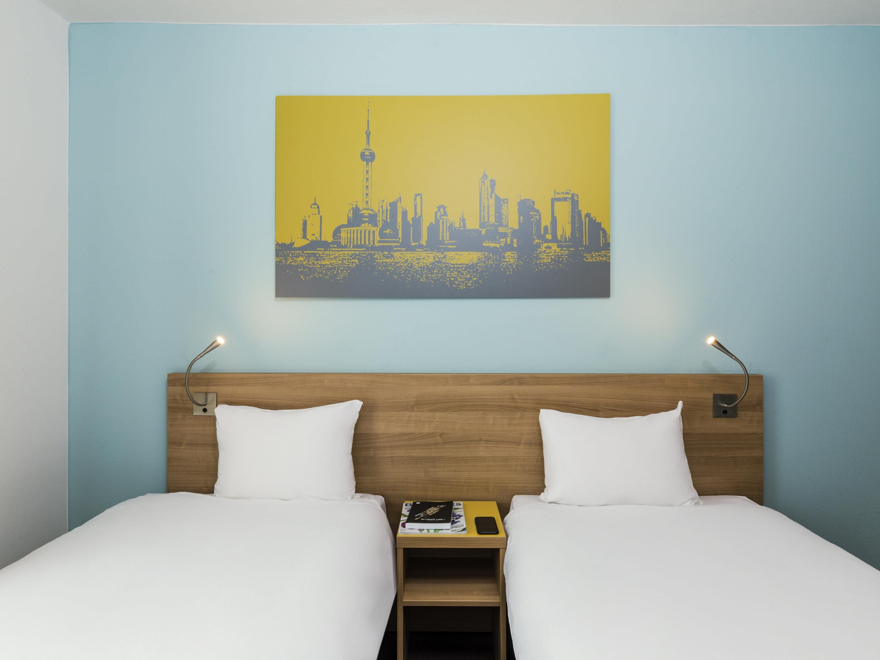 A hotel room with a bed, two pillows, a nightstand with a book and a phone, and a cityscape painting on the wall.