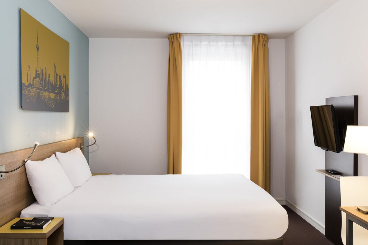 Habitación de hotel con una cama grande, una mesita de noche con un libro, un televisor montado y un cuadro en la pared.