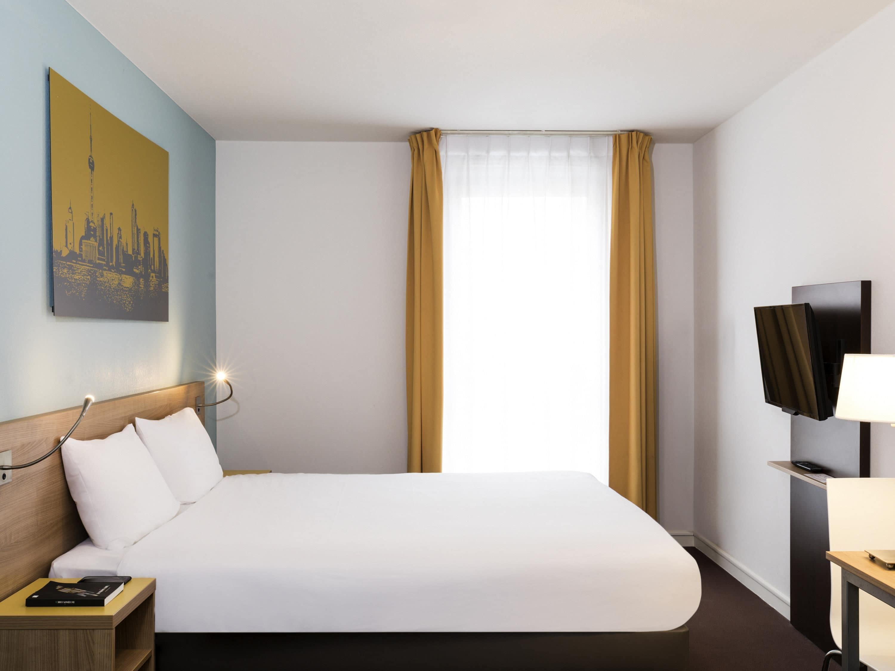 Habitación de hotel con una cama grande, una mesita de noche con un libro, un televisor montado y un cuadro en la pared.