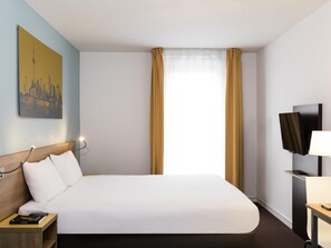 A hotel room with a large bed, a bedside table with a book, a mounted TV, and a painting on the wall.