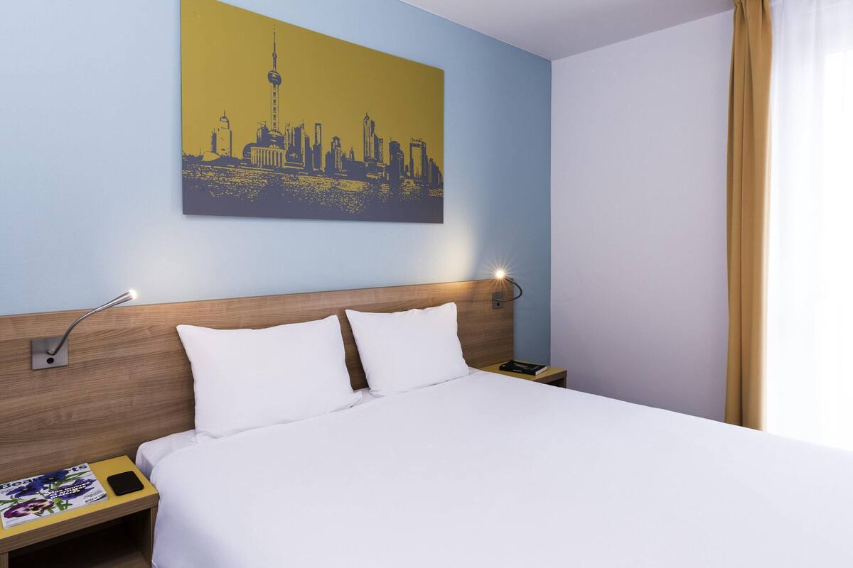 Une chambre d’hôtel avec un lit, une table de chevet, une lampe, un téléphone, un magazine et un tableau représentant un paysage urbain accroché au mur.