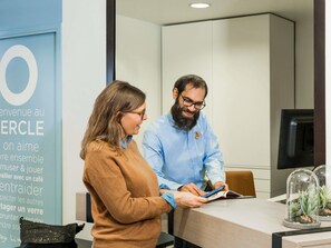 Two people at a reception desk in a modern office setting with a visible wall sign and a small potted plant on the table.