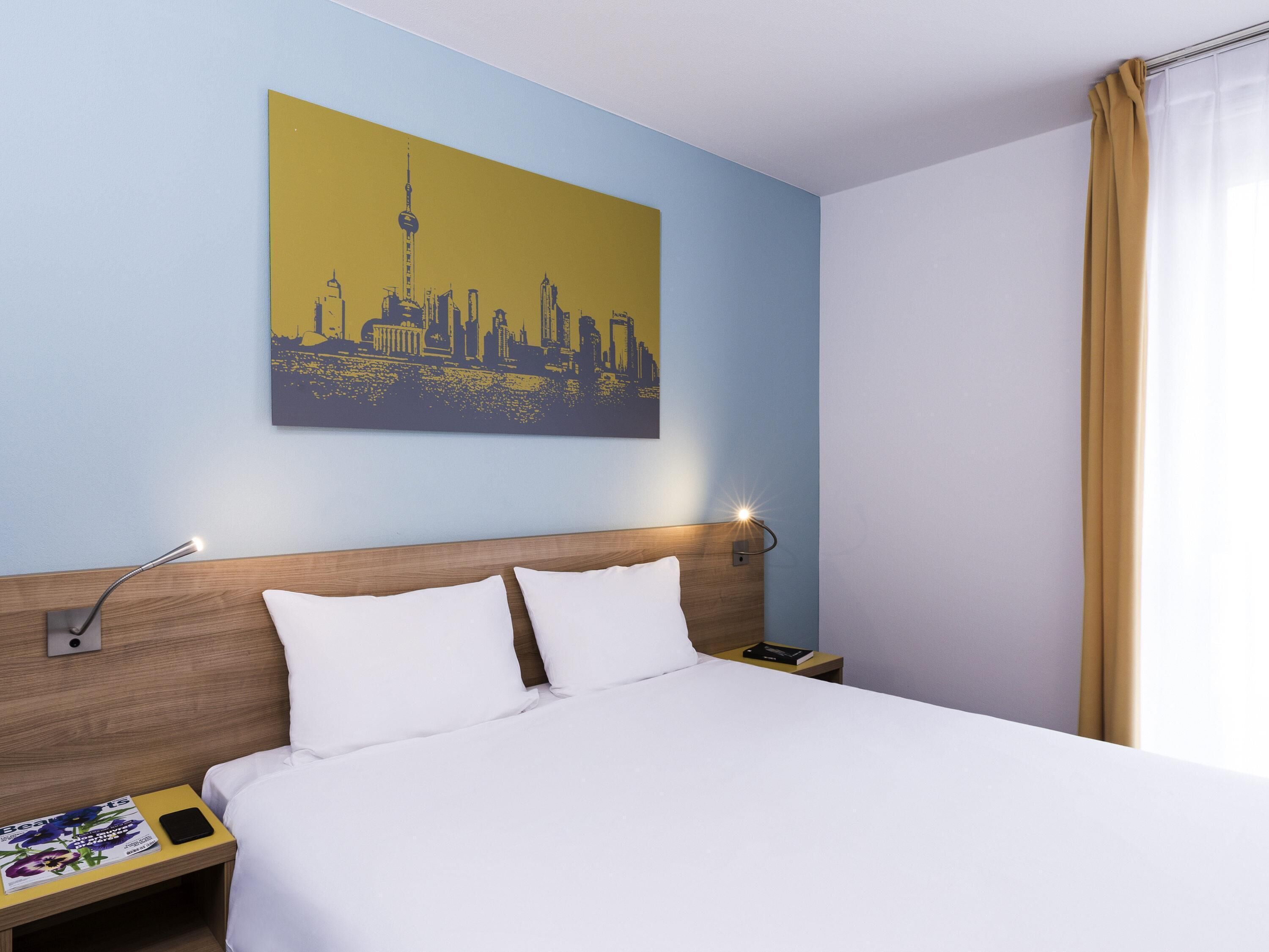 Une chambre d’hôtel avec un lit, une table de chevet, une lampe, un téléphone, un magazine et un tableau représentant un paysage urbain accroché au mur.