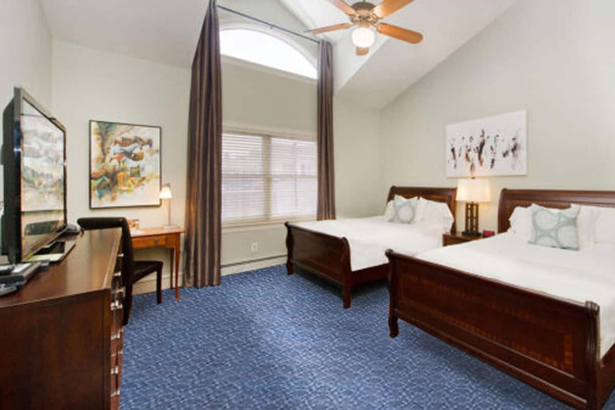 A hotel room with two beds, a desk, a ceiling fan, and artwork on the walls.