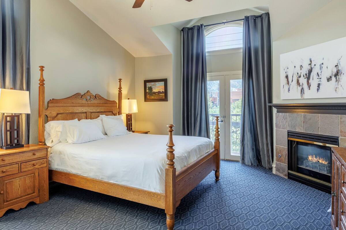A bedroom with a four-poster bed, a nightstand with a lamp, a dresser, a fireplace, and a large window with curtains.
