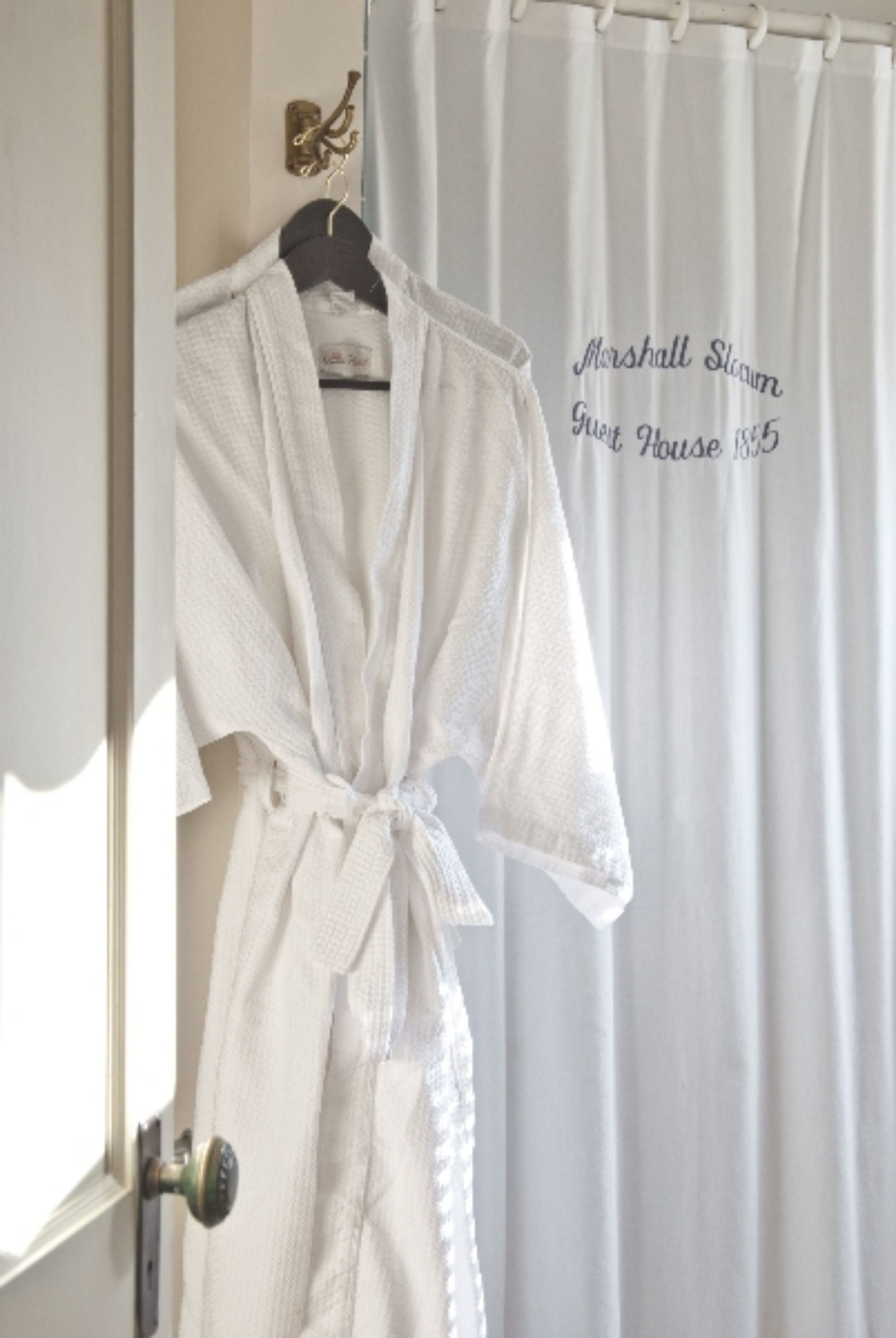 A white robe hanging on a door with a curtain that reads "Marshall Sloan Guest House 1885".