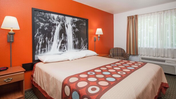 A hotel room with a large bed, two bedside lamps, a chair, a window with curtains, and a wall-mounted air conditioner. - Super 8 by Wyndham Cloverdale Wine Country (Cloverdale)
